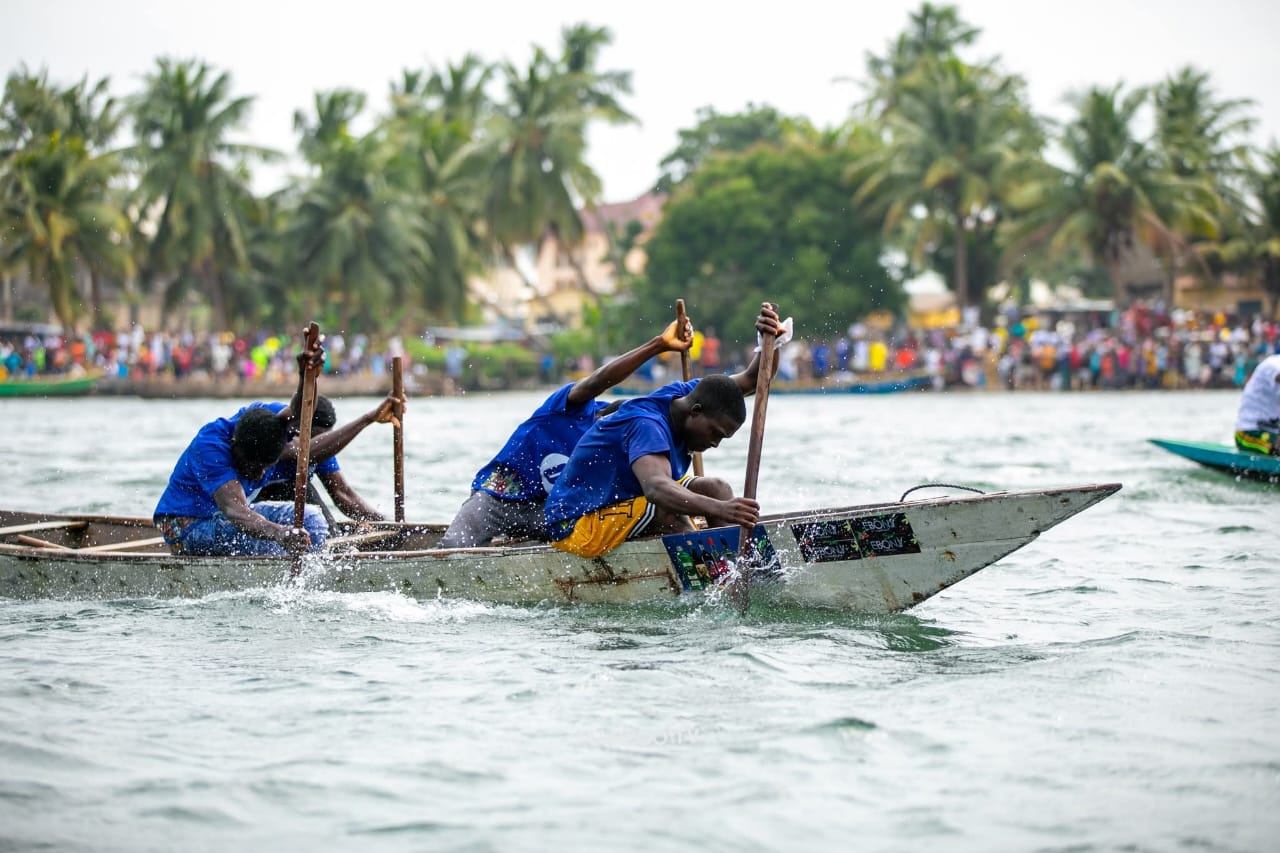 A crew in blue paddles a wooden canoe hard across the water as a crowd watches from the shore